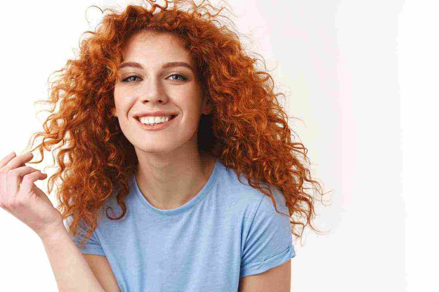 tenderness beauty haircare concept alluring sensual young woman with natural curly red hair rolling strand finger silly smiling toothy looking happy coquettish standing white background
